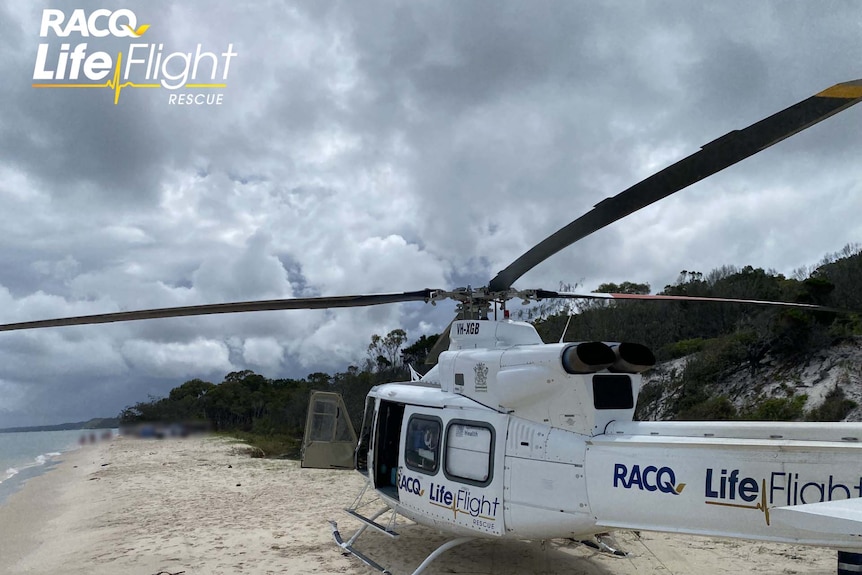 A white RACQ helicopter with its door open is stationary on a beach on a cloudy day.