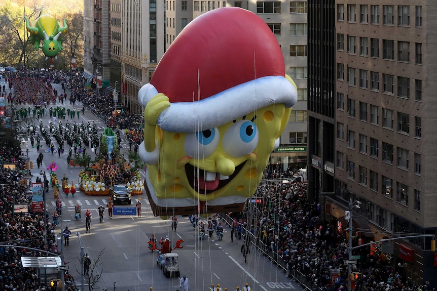 A giant yellow SpongeBob SquarePants balloon with a red and white hat floats above crowds on a New York city street