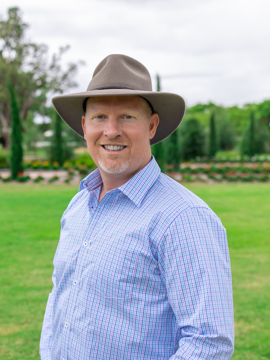  A man with a light brown akubra smiles at the camera before a lush green backdrop.