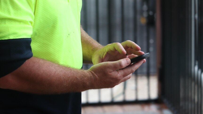 An unidentified man types on his mobile phone, wearing a fluro work shirt.
