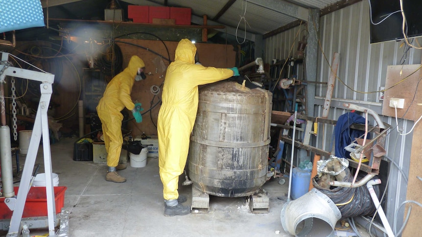 Two people dressed in yellow plastic suits in a shed full of drug paraphernalia