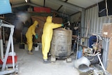 Two people dressed in yellow plastic suits in a shed full of drug paraphernalia