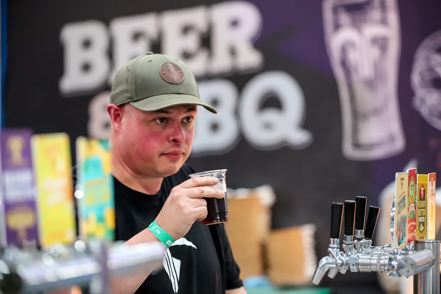 Man wearing cap holds small cup of brown coloured beer, standing behind row of silver beer taps in an exhibitor booth