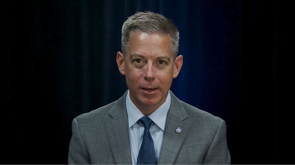 A man with grey hair in a gery suit and navy tie looks serious.