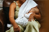 Smiling woman breastfeeding her baby on a brown leather couch. She has a white muslin draped over her shoulder.