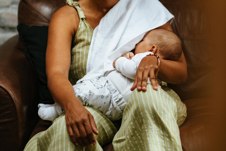 Smiling woman breastfeeding her baby on a brown leather couch. She has a white muslin draped over her shoulder.