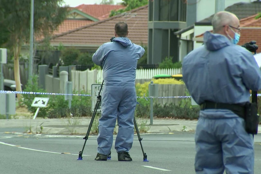 Two forensic police officers are wearing blue plastic suits and face masks as they photograph the taped-off crime scene.