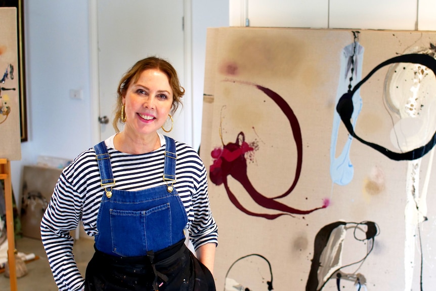 A woman in striped top and overalls stands in front of an art work