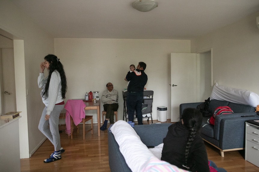A woman with long black hair walks out of a room, while a man holds a baby, older man sits on chair, a woman's back to camera.