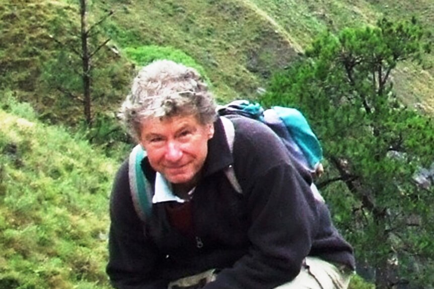 A man with grey hair and a backpack stands in green hills.