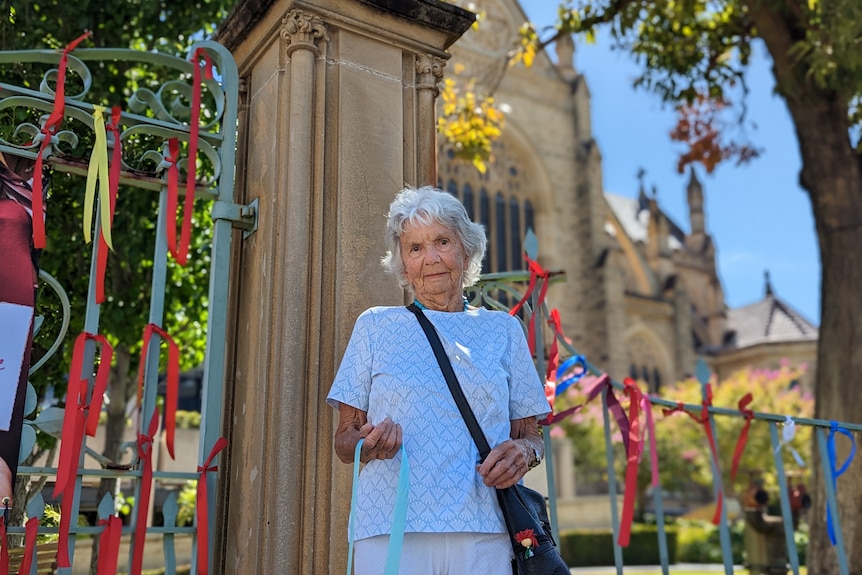Pam Day stands holding a ribbon with the church in the background and ribbons hanging on the fence around her. 