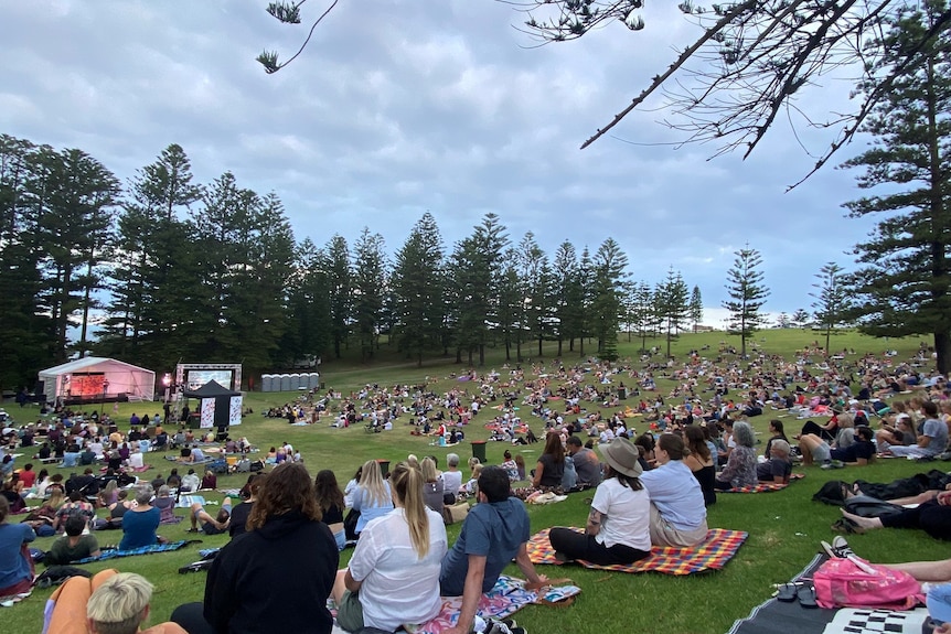 Hundreds of people sitting on rugs in a large park watching performers on a stage.