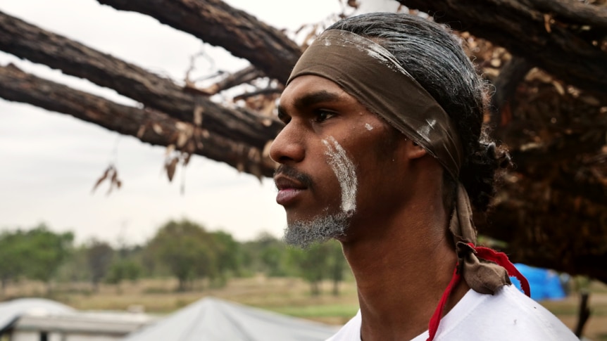 a man in cultural face paintings and a white shirt looks off into the distance