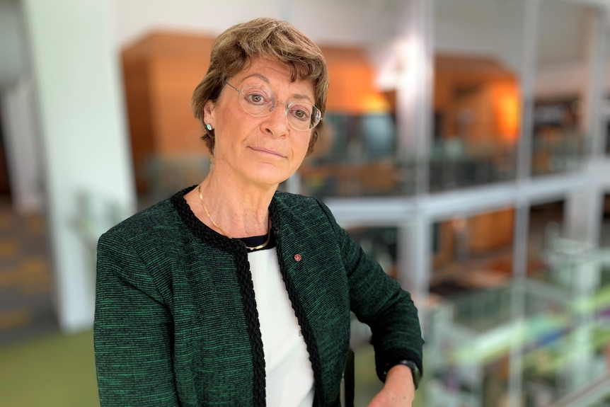 a woman with short hair and glasses wearing a dark green jacket.