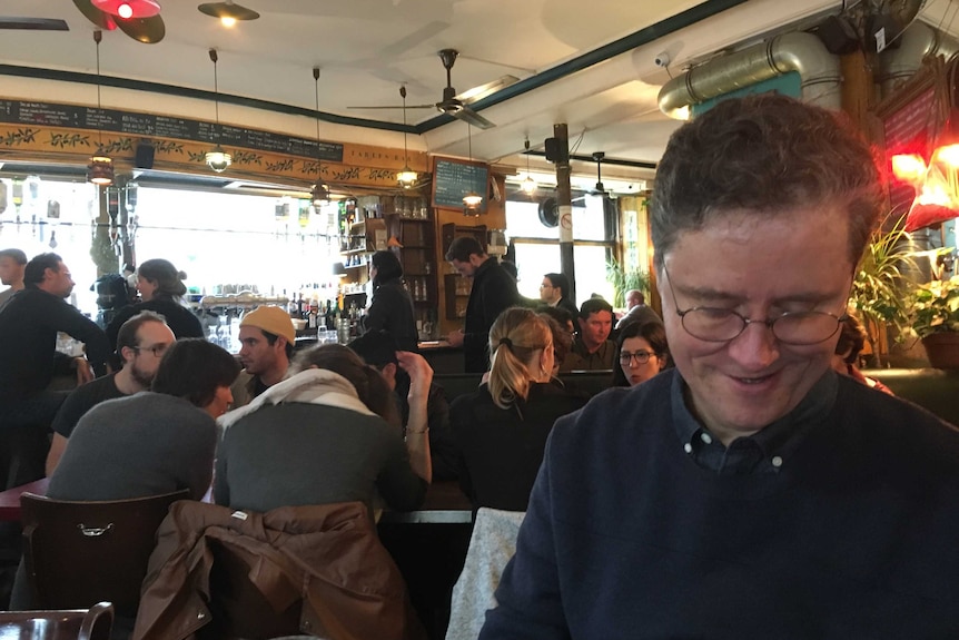 A man wearing glasses and a black shirt sitting inside a cafe and reading.