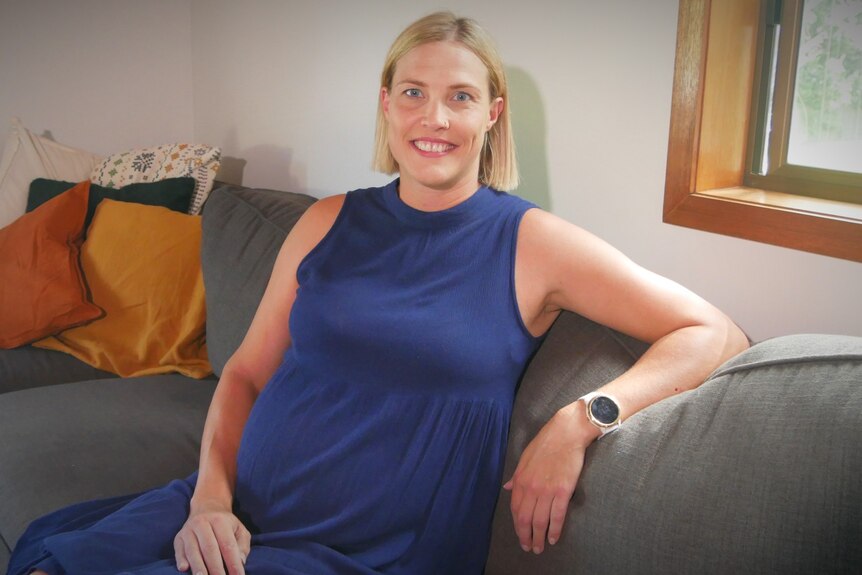 A blonde pregnant woman sitting on a couch smiling at the camera