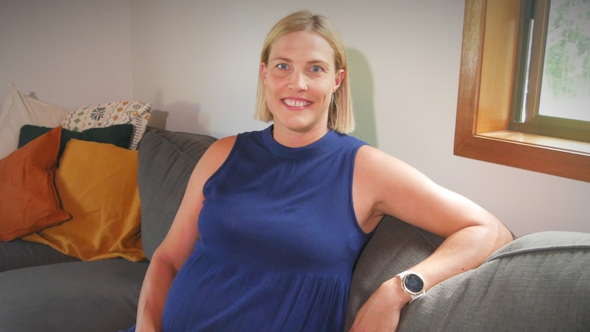 A smiling blonde woman, who is pregnant, sits on a couch.