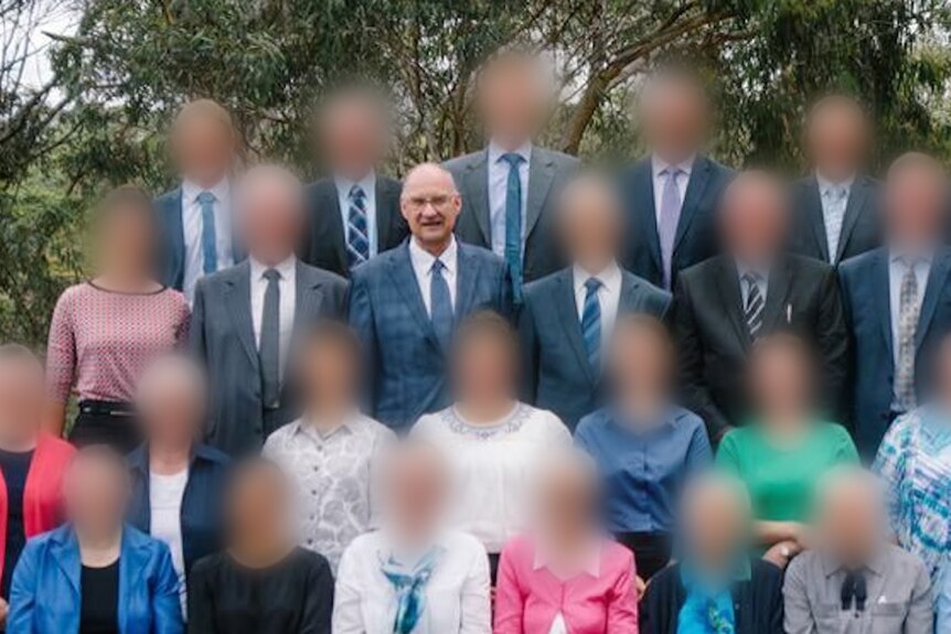 A group of people lined up like a school photo with their faces blurred. One elderly man has his face unblurred