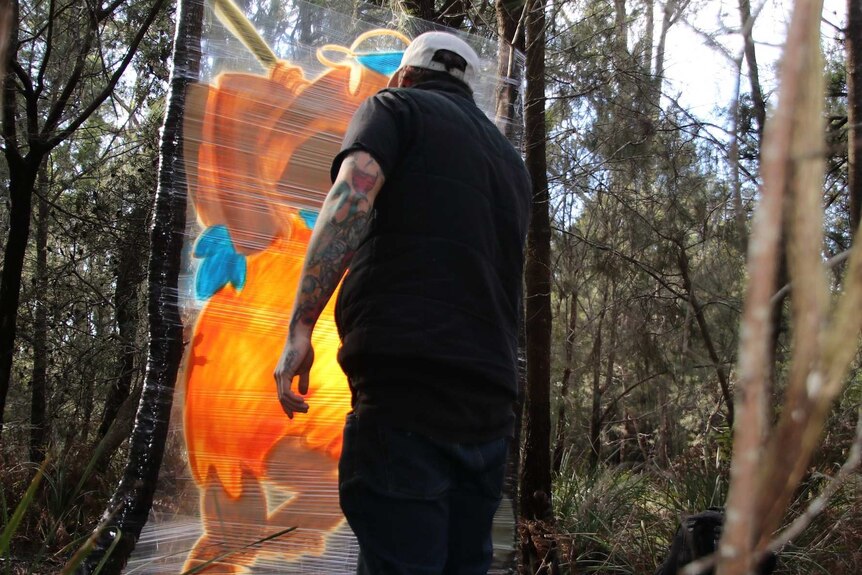 Tasmanian artist James Cowan spray painting a mural on plastic sheeting stretched between trees.