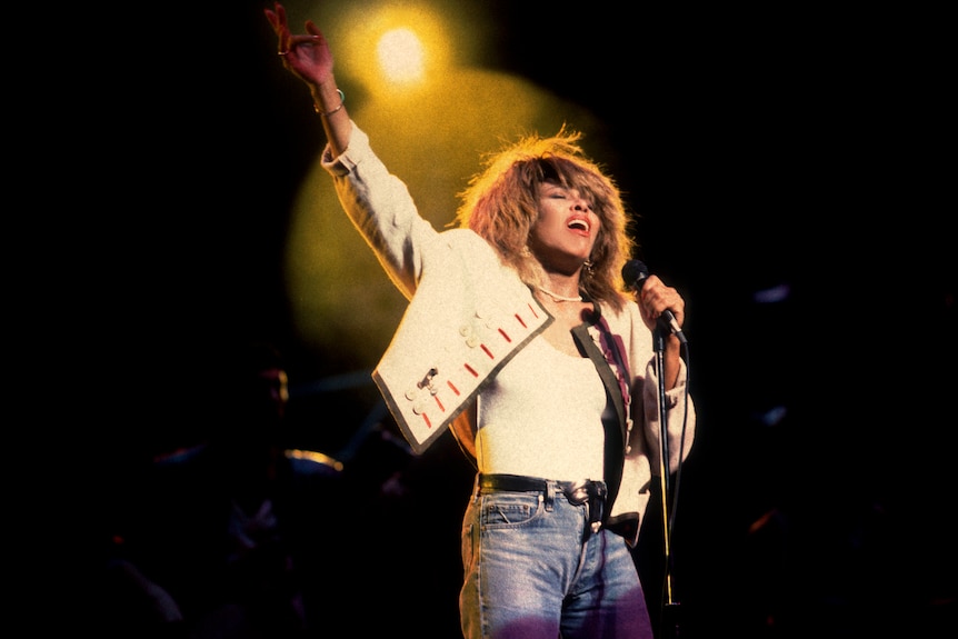 Tina Turner, a Black woman with voluminous blonde hair wearing a white jacket and blue jeans performs under yellow light onstage