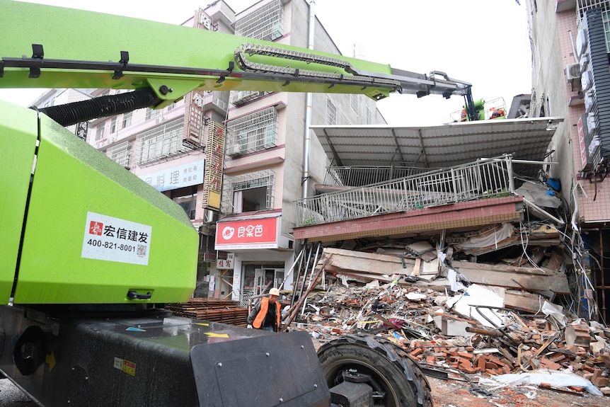 A large crane operates on a collapsed building, which sits demolished between two standing buildings