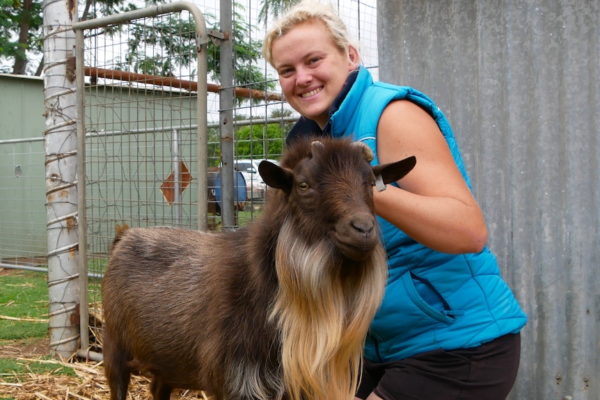 A young woman grins holding onto a small male goat with a dark shaggy coat and a long blonde beard