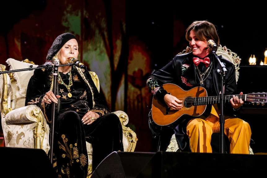 Joni Mitchell and Brandi Carlile performing on stage, Carlile holding a guitar. Mitchell dressed in black