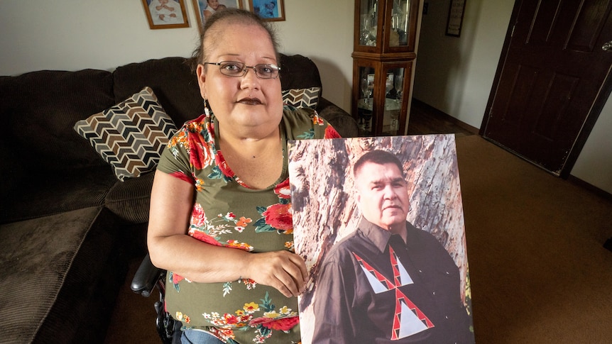 Woman sitting in a chair holding up a large portrait of her husband. 