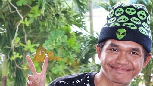 A man with a fun hat and drink next to him sits making a V (victory) sign with his fingers.