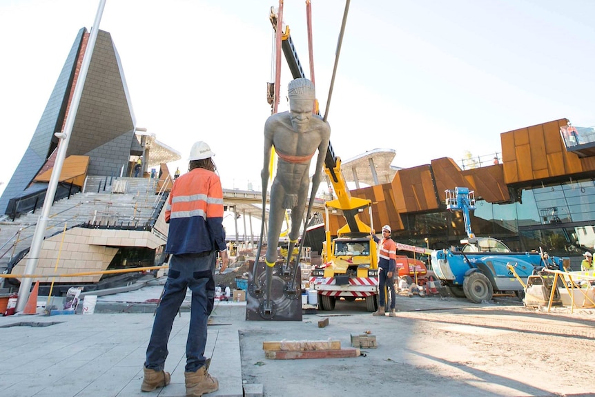 A tall grey statue is pulled upright with a crane as a worker looks on with the buildings of Yagan Square in the background.