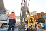 A tall grey statue is pulled upright with a crane as a worker looks on with the buildings of Yagan Square in the background.