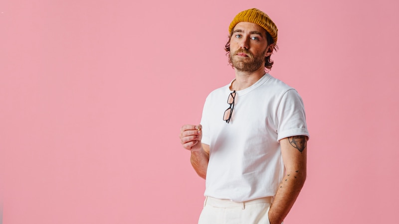 Matt Corby wearing a white t-shirt and coral beanie against a light pink backdrop