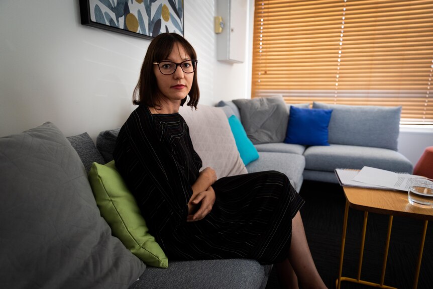 A woman with a neutral expression wearing glasses, sitting on a couch in an office.