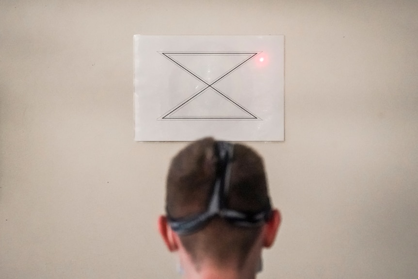 The back of a teenage boy's head, with a laser being pointed at a shape on a sheet of paper stuck to a wall.