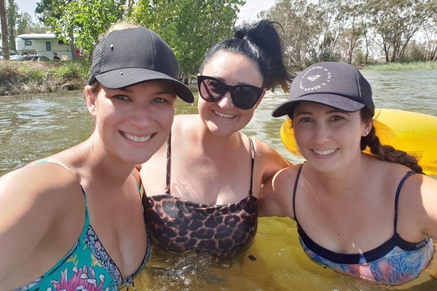 Three women in a lake smile at a camera.