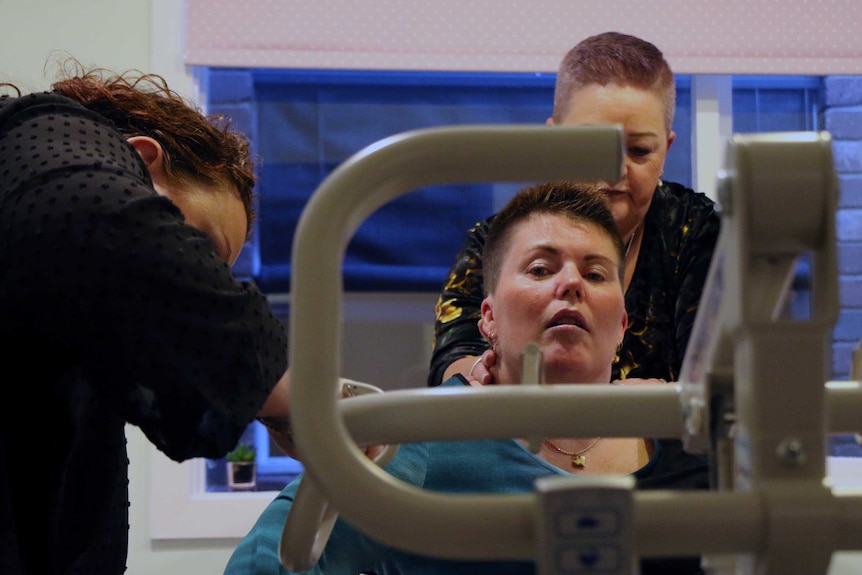 Sarah Brady looks at the camera as her carer and mother help her out of bed with a mechanical aid.