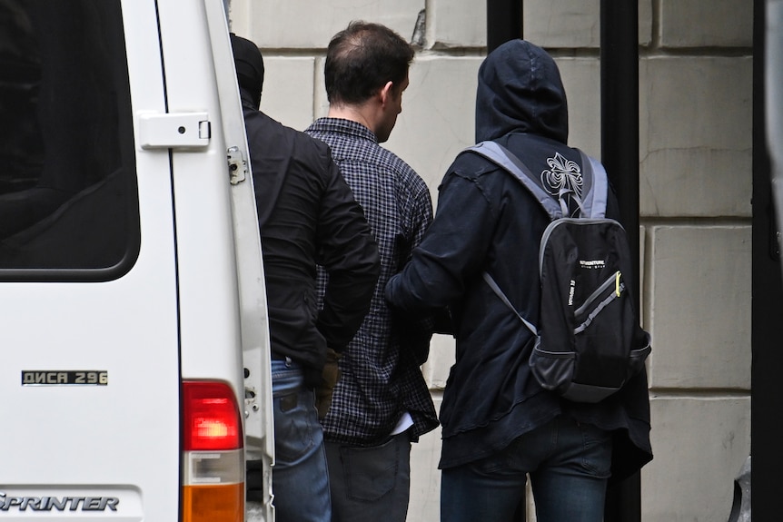 Two people wearing hooded jumpers leading another man by the arm from a white van towards a building