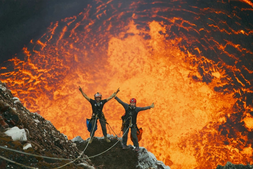 Two men wearing harnesses stand on the rocky edge of a volcano with bright burning lava behind them.