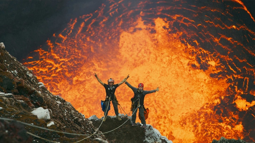 Two men stand with harnesses on the rocky edge of a volcano with bright burning lava behind them