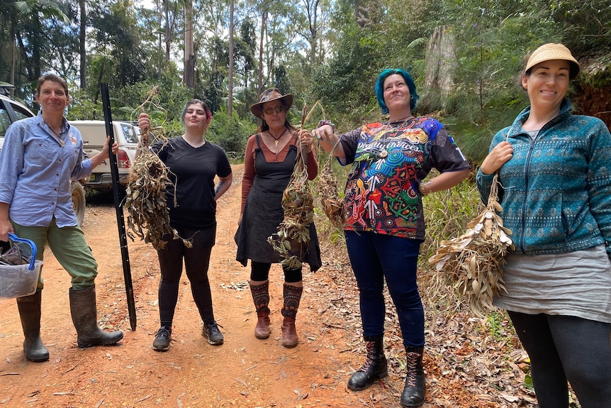 Four women and a man stand on a forest road holding woven nests, all smiling, red dirt, some wear hats, one carries black stick.