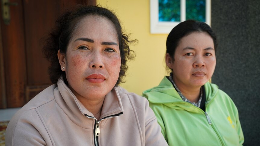 Two women sit side by side in front of a yellow wall. One wears beige zip-up jumper, the other lime-green. Both look serious
