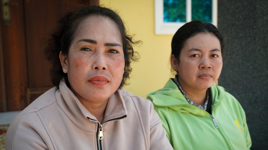Two women sit side by side in front of a yellow wall. One wears beige zip-up jumper, the other lime-green. Both look serious