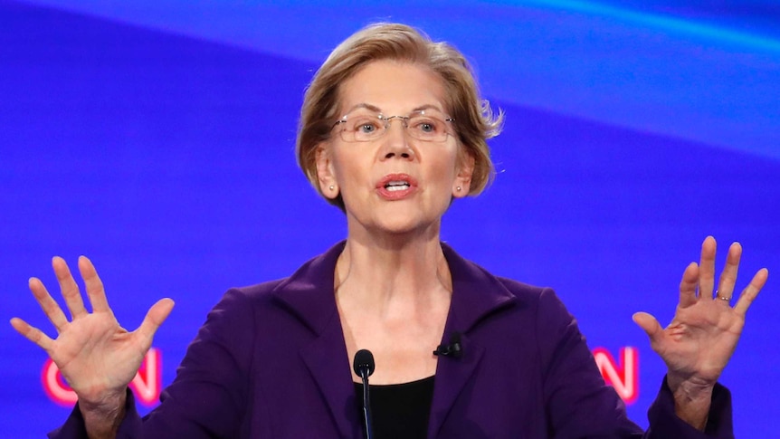 Elizabeth Warren stands at a lectern with her palms facing outwards, in front of a wall with CNN logos