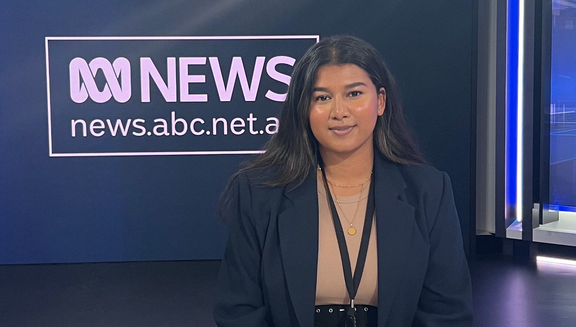Young woman with dark hair and tanned skin sitting behind a news desk. She is wearing a tan shirt and dark blue blazer
