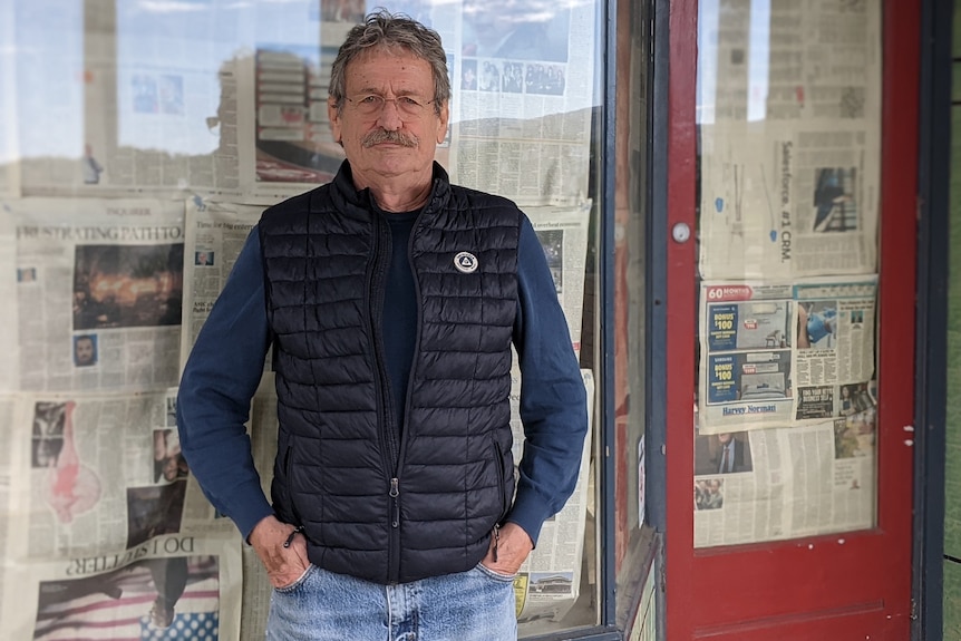 A man wearing a black puffer vest and blue shift stands in front of shop windows covered in newspaper.