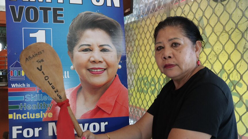 Woman holds a wooden spoon in front of a political corflute poster of her face