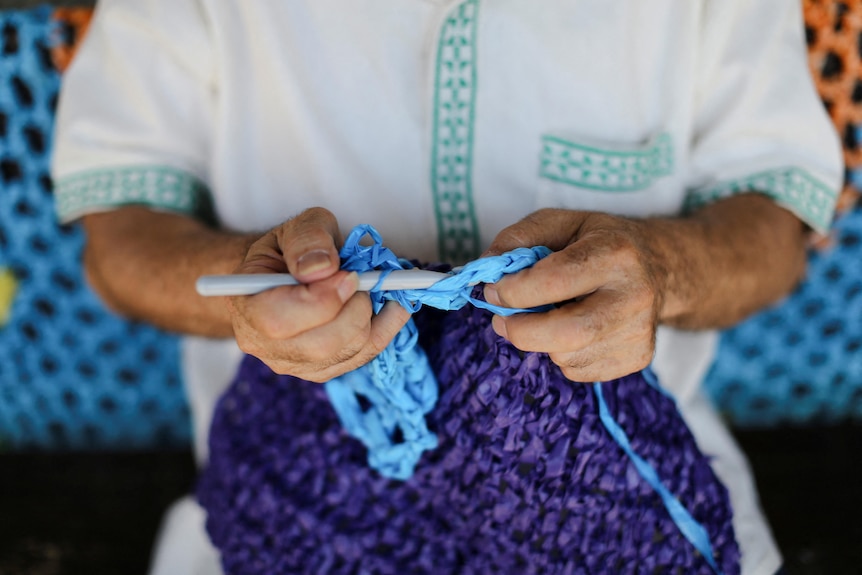 A man's hands are weaving a blue canopy using a knitting needle. 