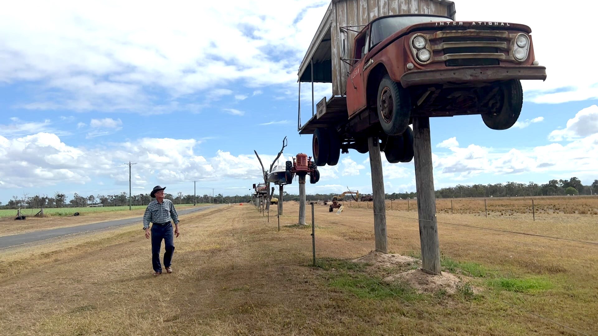 Elderly man walking beside a road, looking at a row of old vehicles on top of old power poles.