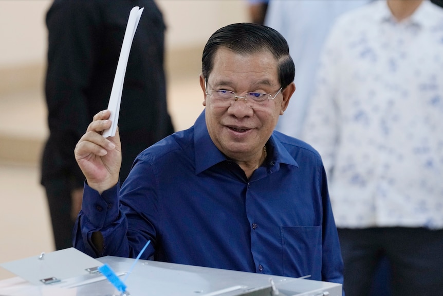 Hun Sen is wearing a blue business shirt holding up a ballot paper before placing it into a box. 