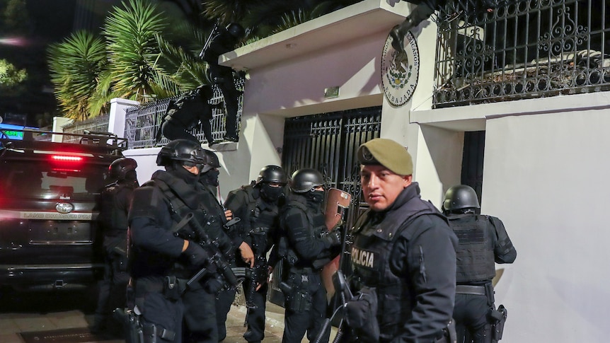 A group of heavily armed police in dark blue uniforms stand outside and scale a spiked wall at night.